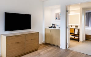 A hotel room with a flat-screen TV mounted on the wall, a wooden cabinet, a kitchenette with a coffee machine, and a bathroom visible through an open door.