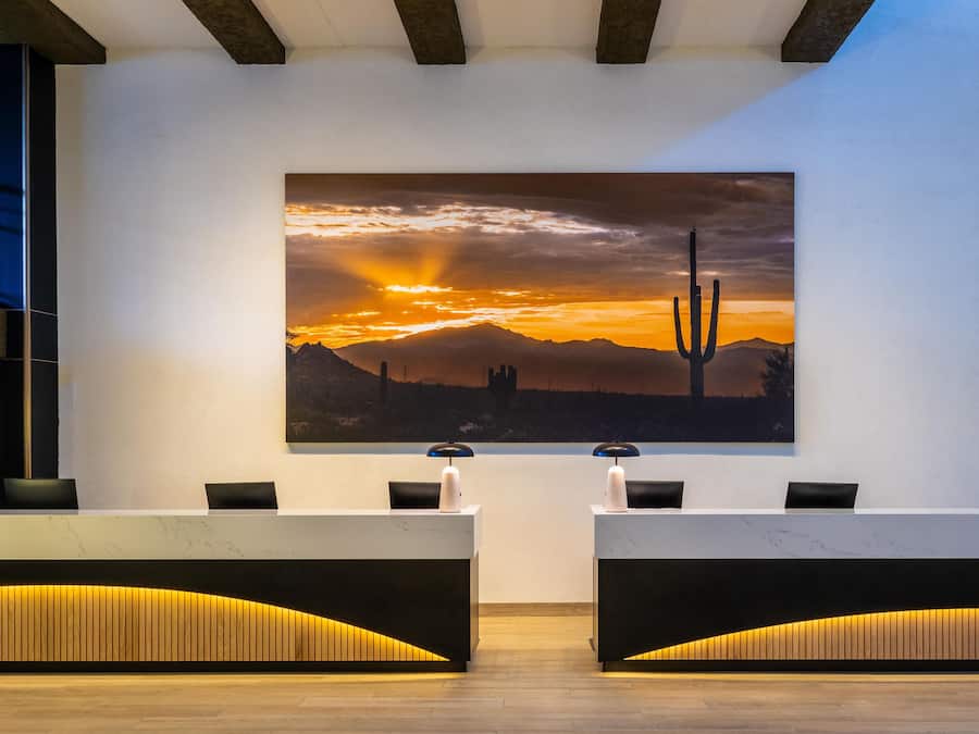 Zona de recepción con un cuadro grande de atardecer desértico, dos mostradores de recepción y un diseño moderno con detalles de madera.