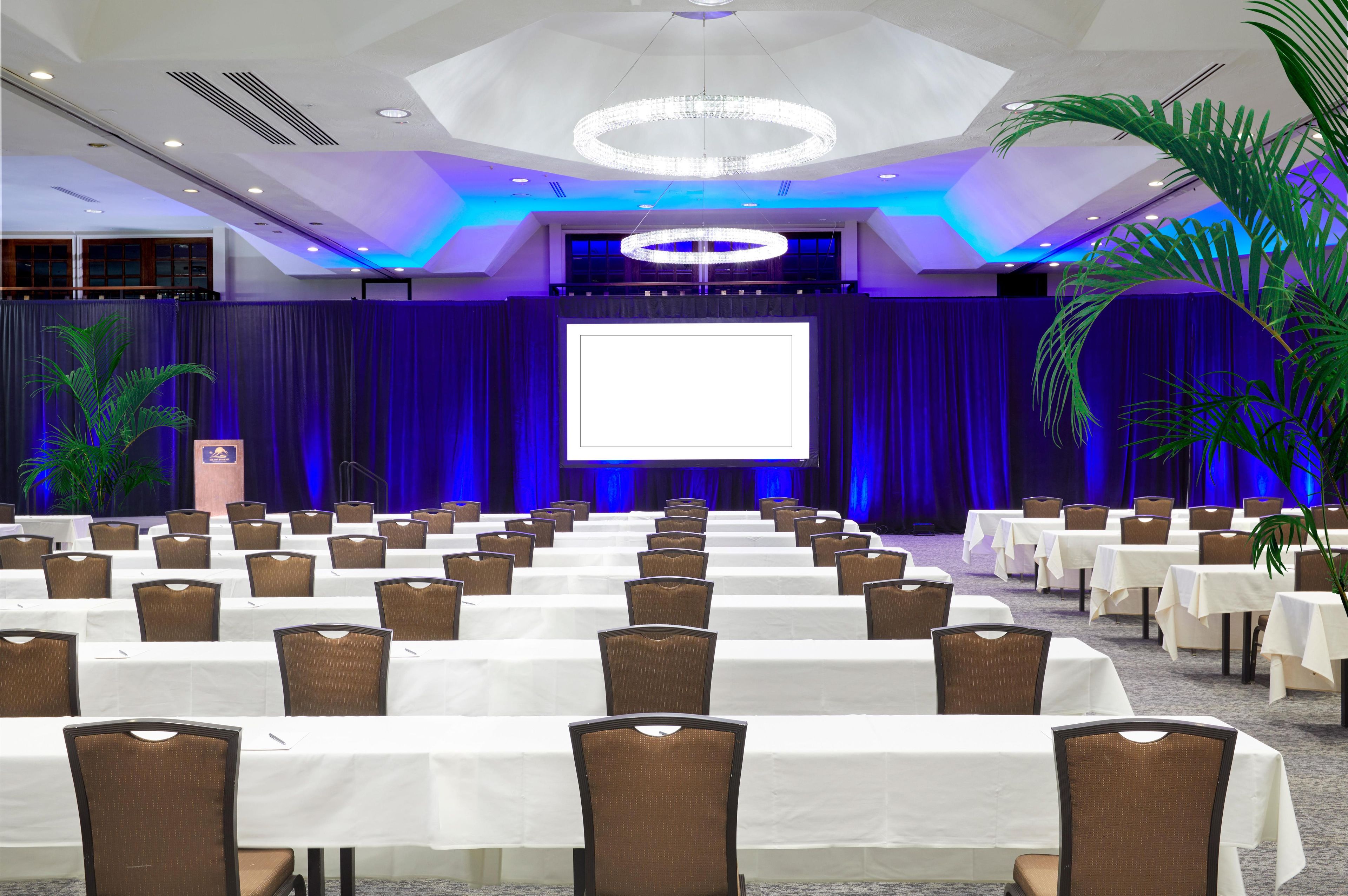 A conference room with rows of tables and chairs, a large screen at the front, and a stage with curtains.