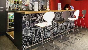 A bar area with a black and white mural, a refrigerator with beverages, and modern bar stools. - ibis Paris Pont De Suresnes (Suresnes)
