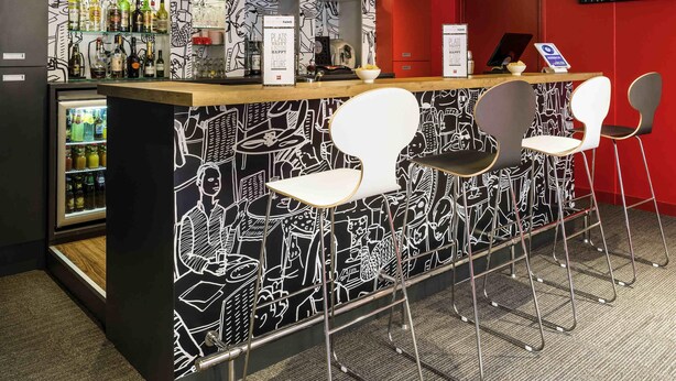 A bar area with a black and white mural, a refrigerator with beverages, and modern bar stools. - ibis Paris Pont De Suresnes (Suresnes)