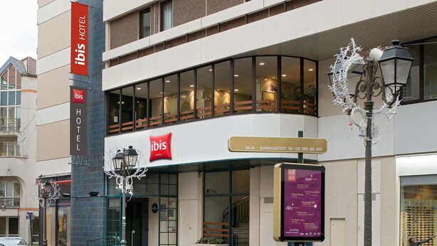 A modern hotel with a prominent 'ibis' sign, a glass entrance, and a street lamp with decorative lights. - ibis Paris Pont De Suresnes (Suresnes)