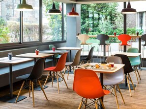 A modern cafe interior with tables and chairs, large windows with a view of greenery, and pendant lighting. - ibis Paris Pont De Suresnes (Suresnes)