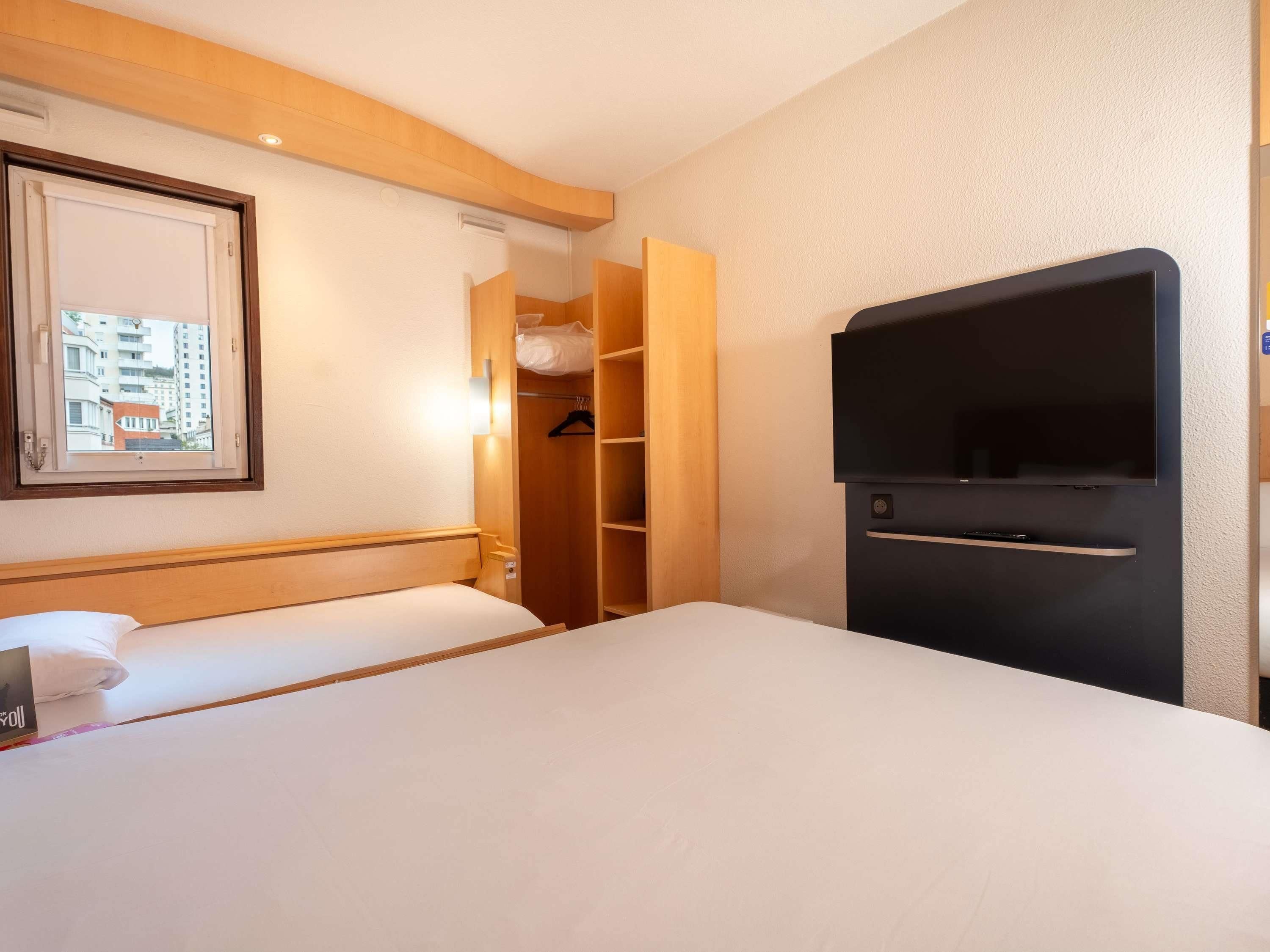A hotel room with a bed, a TV, a wardrobe, and a window with a city view.
