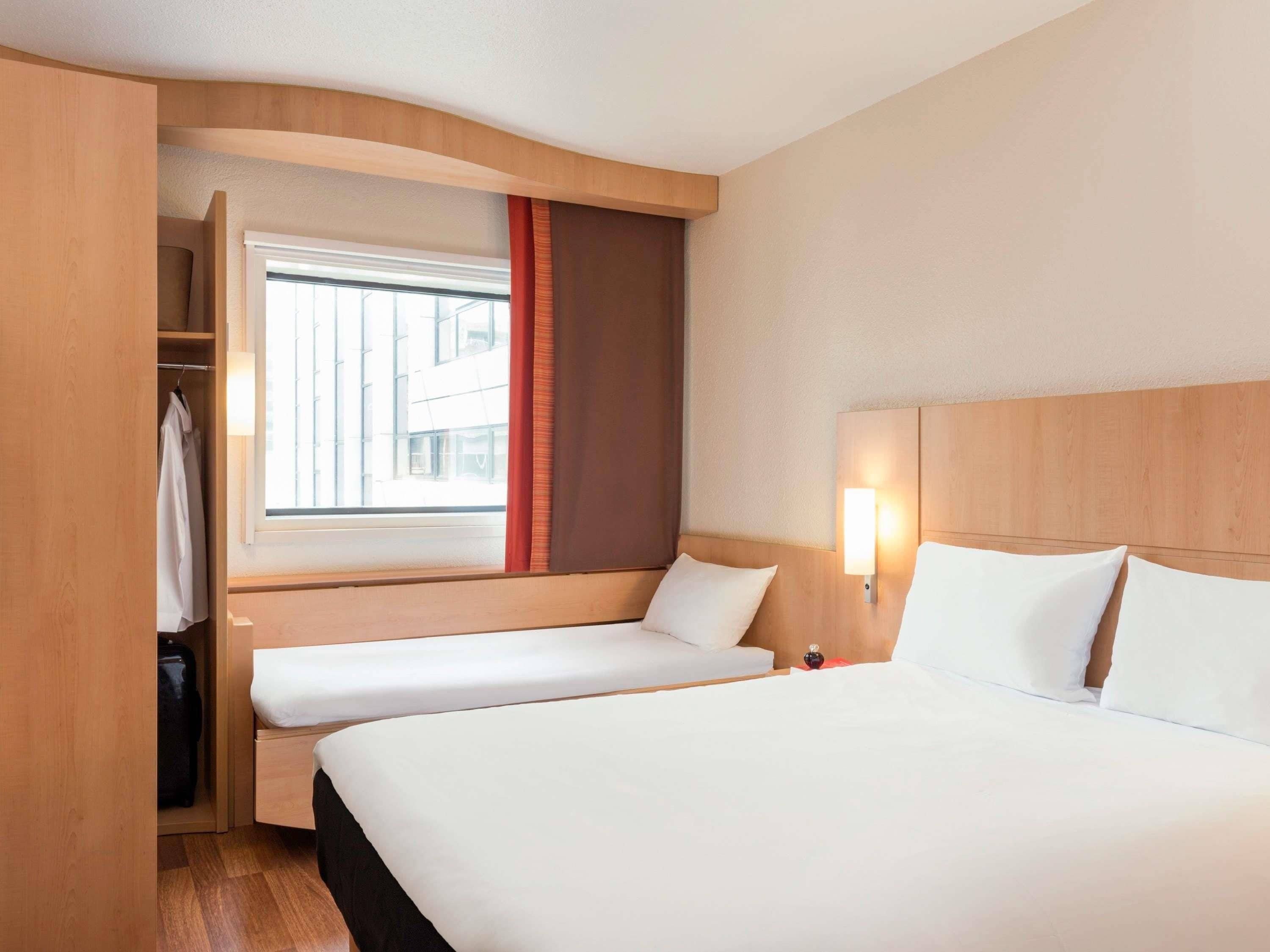 Une chambre d’hôtel avec un grand lit, un lit plus petit, une fenêtre avec des rideaux, une armoire et une lampe de chevet.