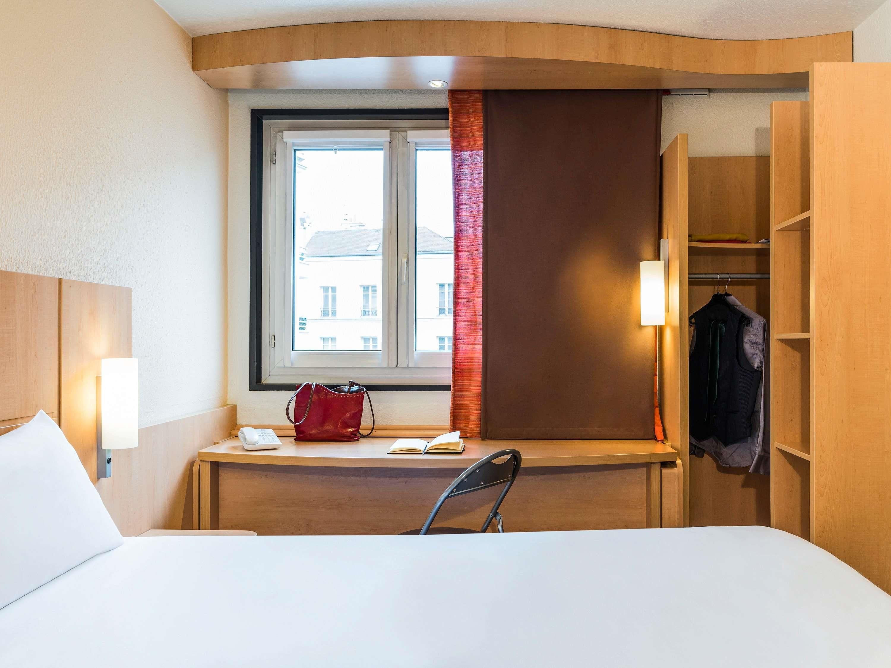Ett hotellrum med en säng, ett skrivbord med en stol, ett fönster med gardiner, en sänglampa, en klädställning och en garderob.