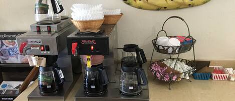 A coffee station with multiple coffee makers, a basket, and a tray with cups and utensils.