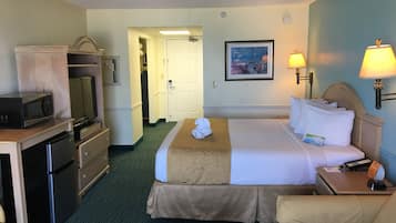A hotel room with a bed, a desk with a microwave, a refrigerator, a chair, and a nightstand with a lamp.