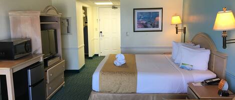A hotel room with a bed, a desk with a microwave, a refrigerator, a chair, and a nightstand with a lamp.