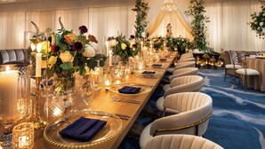 Une longue table de banquet, dressée avec de la belle porcelaine, de la verrerie raffinée et d’élégantes serviettes, entourée de fauteuils confortables dans une pièce bien éclairée, où trône un lustre.