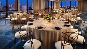 Une salle de banquet avec des tables rondes disposées pour un événement formel, de grandes fenêtres offrant une vue sur la ville et un sol recouvert de moquette.