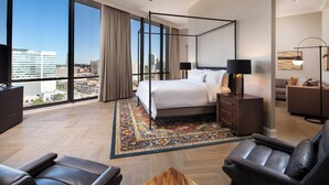 Une chambre d’hôtel moderne avec un lit à baldaquin, une grande fenêtre donnant sur la ville, un canapé en cuir et une table basse en bois.