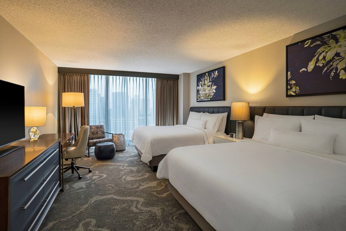 Een hotelkamer met twee bedden, een bureau met een televisie, een stoel en een groot raam met gordijnen.