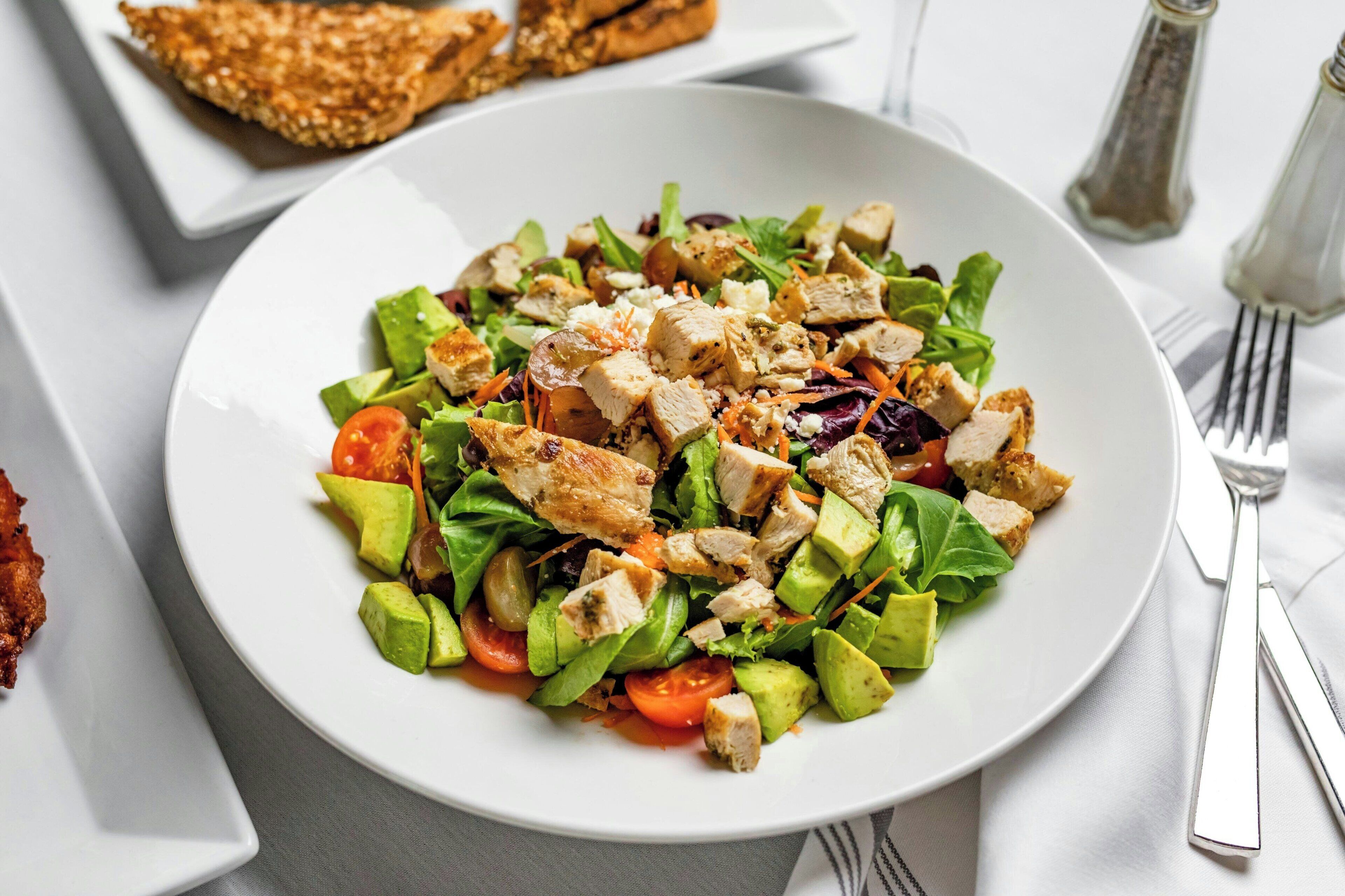 A plate of salad with avocado, tomatoes, and chicken on a white plate.