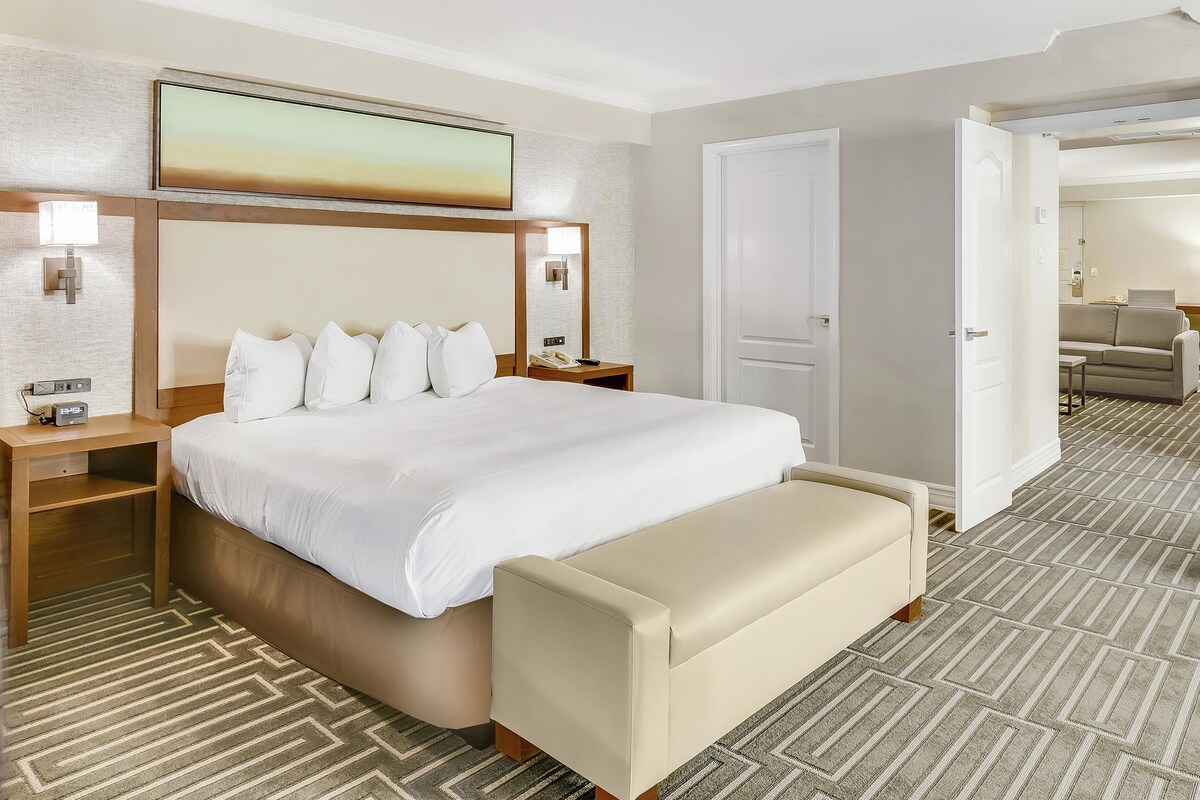 A hotel room with a large bed, bedside tables, a bench, a painting, and a patterned carpet.