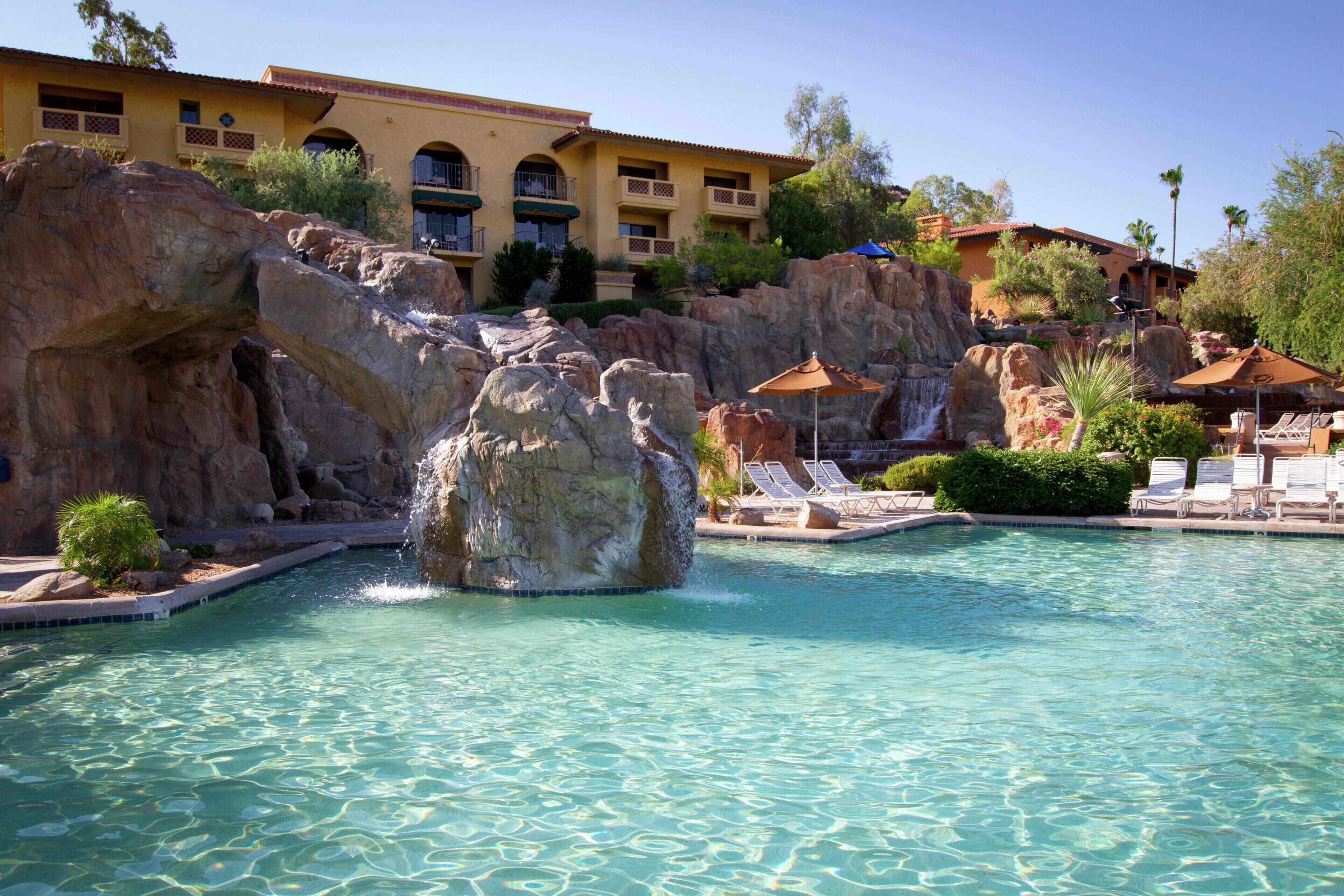 A pool with a rock waterfall, lounge chairs, and a building with balconies.