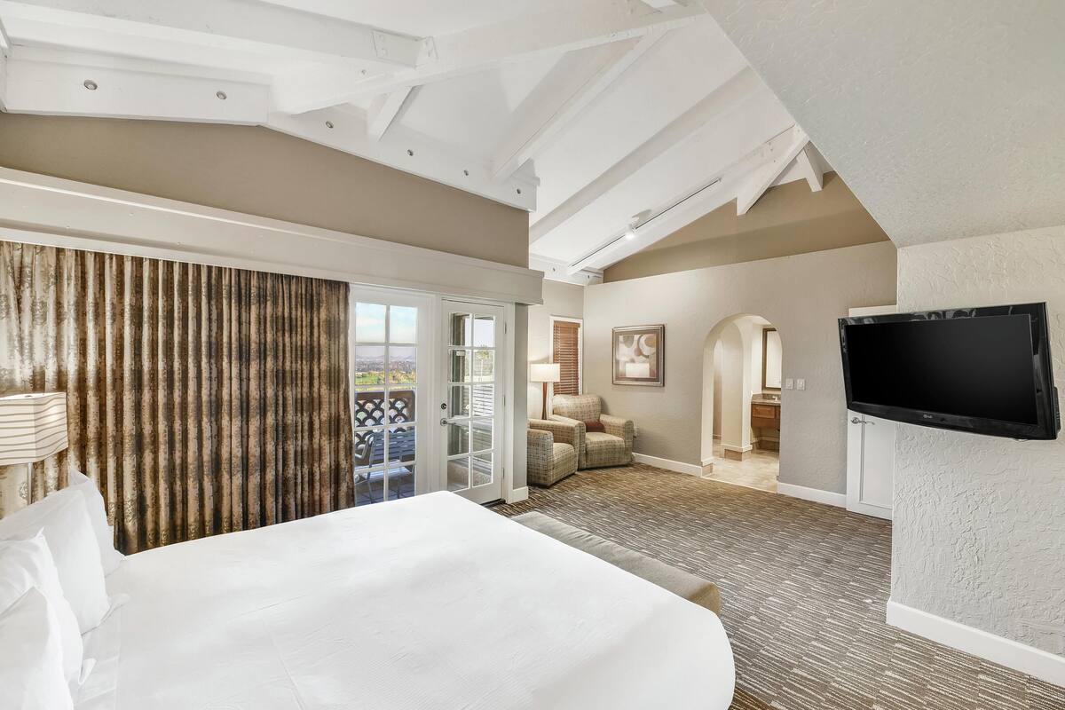 Habitación de hotel con una cama grande, televisión, puerta corrediza a un balcón y piso alfombrado.