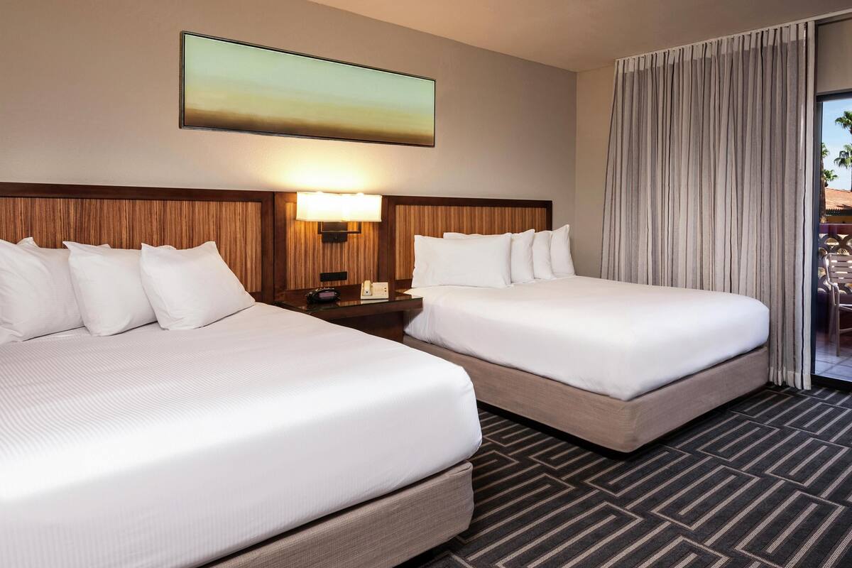 Una habitación de hotel con dos camas, una mesita de noche, una lámpara y un ventanal con cortinas.