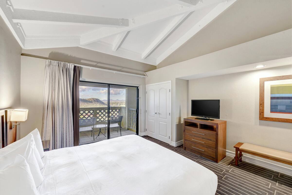 Habitación de hotel con una cama grande, televisión, cómoda de madera, banco y balcón con vistas.