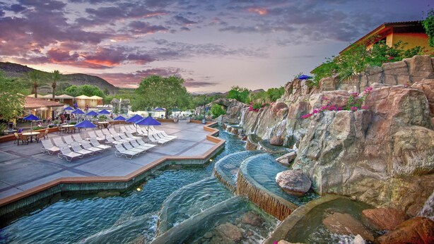 A pool area with lounge chairs and umbrellas, a rocky landscape, and a sunset sky.