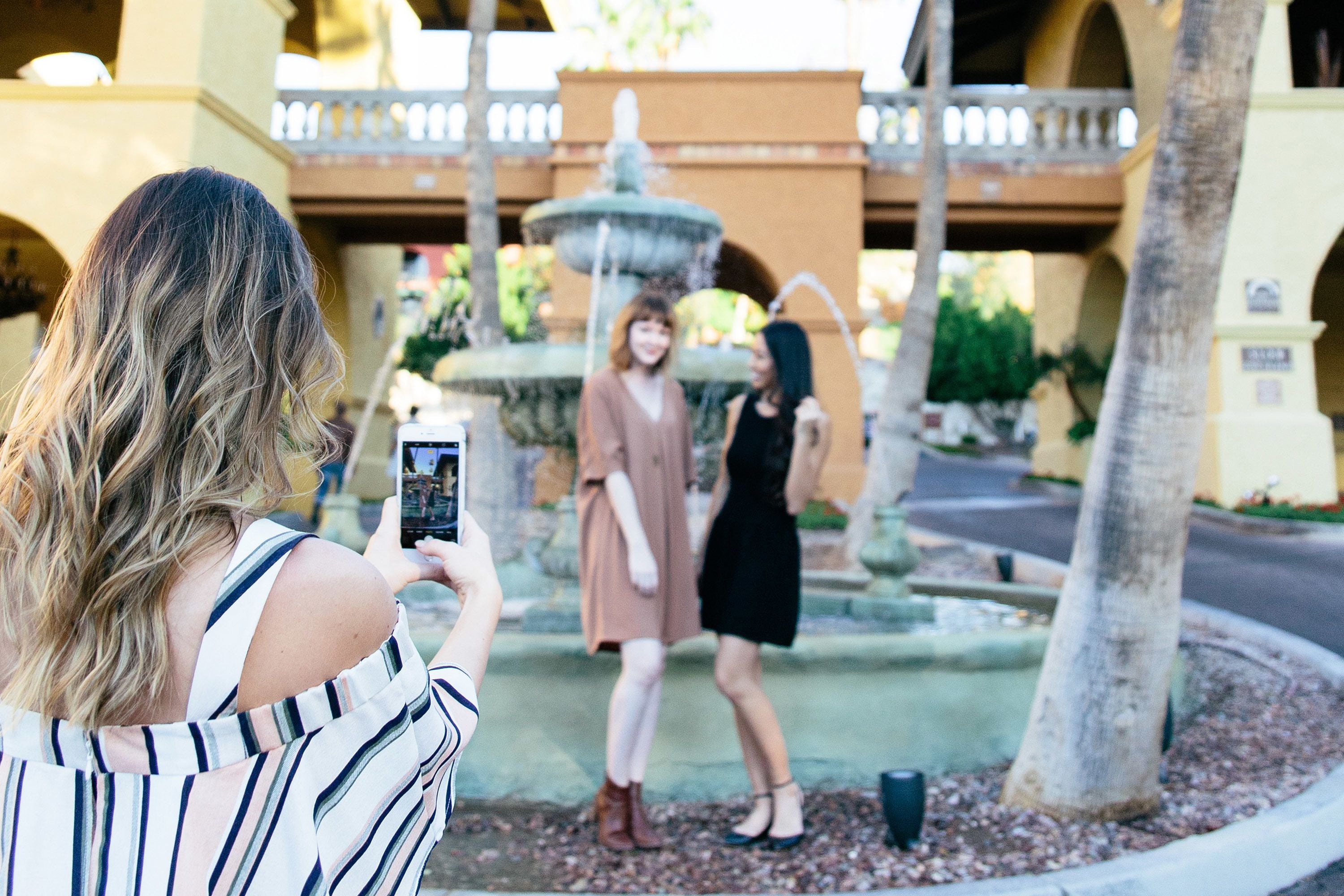 A person taking a photo of two individuals standing in front of a fountain in an outdoor setting with a building in the background.