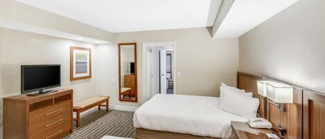 Habitación de hotel con cama, televisor, escritorio, espejo y puerta que da a un balcón.