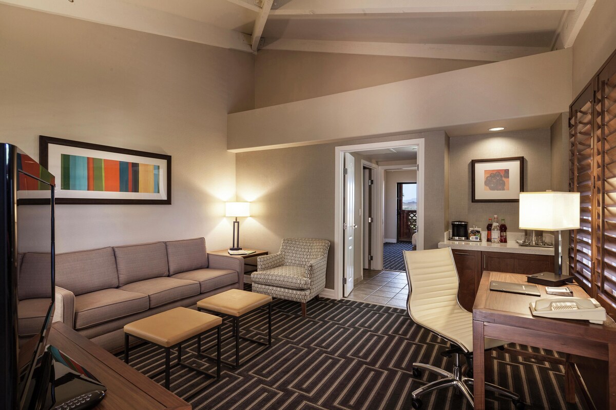 A hotel room with a sofa, armchair, desk, and a striped carpet.