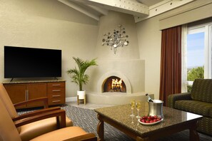A hotel room with a flat-screen TV, a fireplace, a dining table with a fruit bowl, and a sofa.