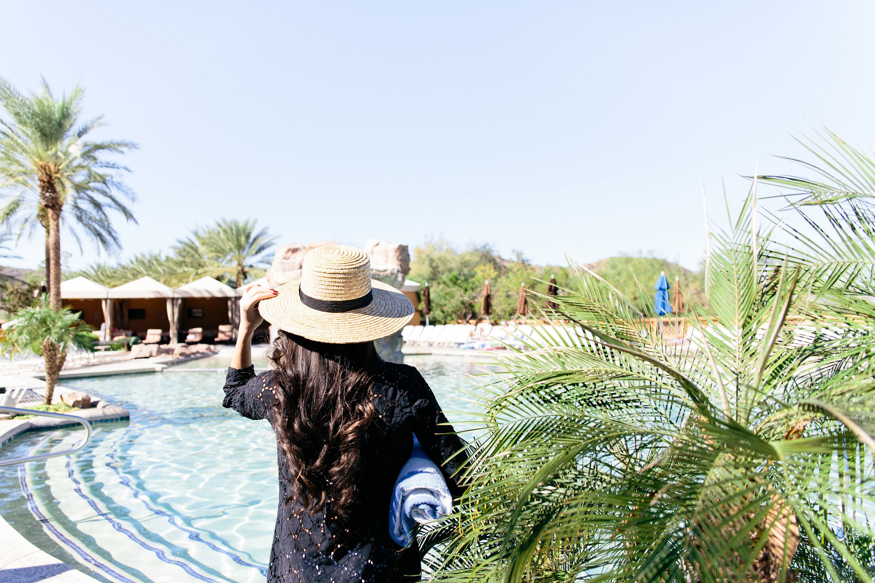 Una persona de pie junto a una piscina, con palmeras y sillas de descanso al fondo.