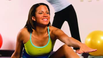 A person sitting on a blue exercise mat in a room with another person standing in the background.