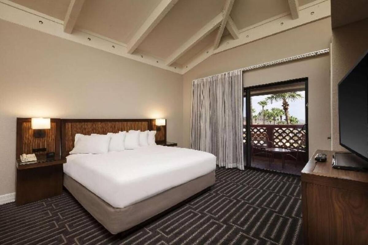 A hotel room with a large bed, a TV, a nightstand, and a view through a door to an outdoor area with a balcony.