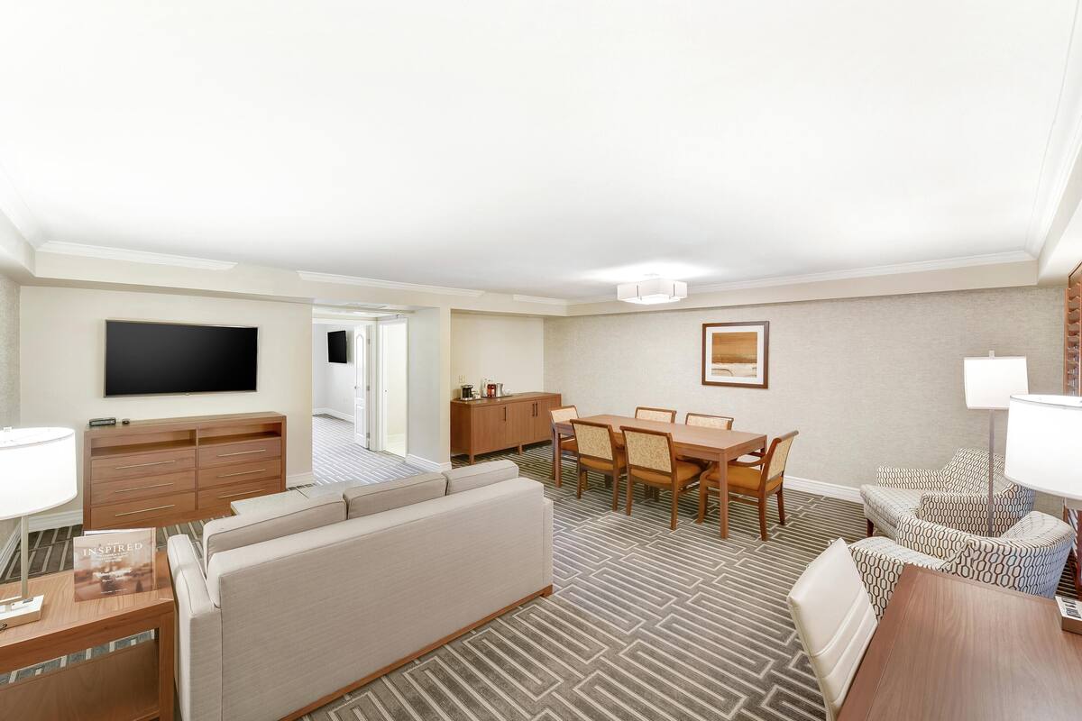 Habitación de hotel con televisor de pantalla plana, sofá, mesa de comedor con sillas, alfombra estampada y un armario.