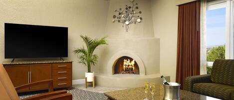 A hotel room with a flat-screen TV, a fireplace, a dining table with a bottle of champagne, and a view of the outdoors.
