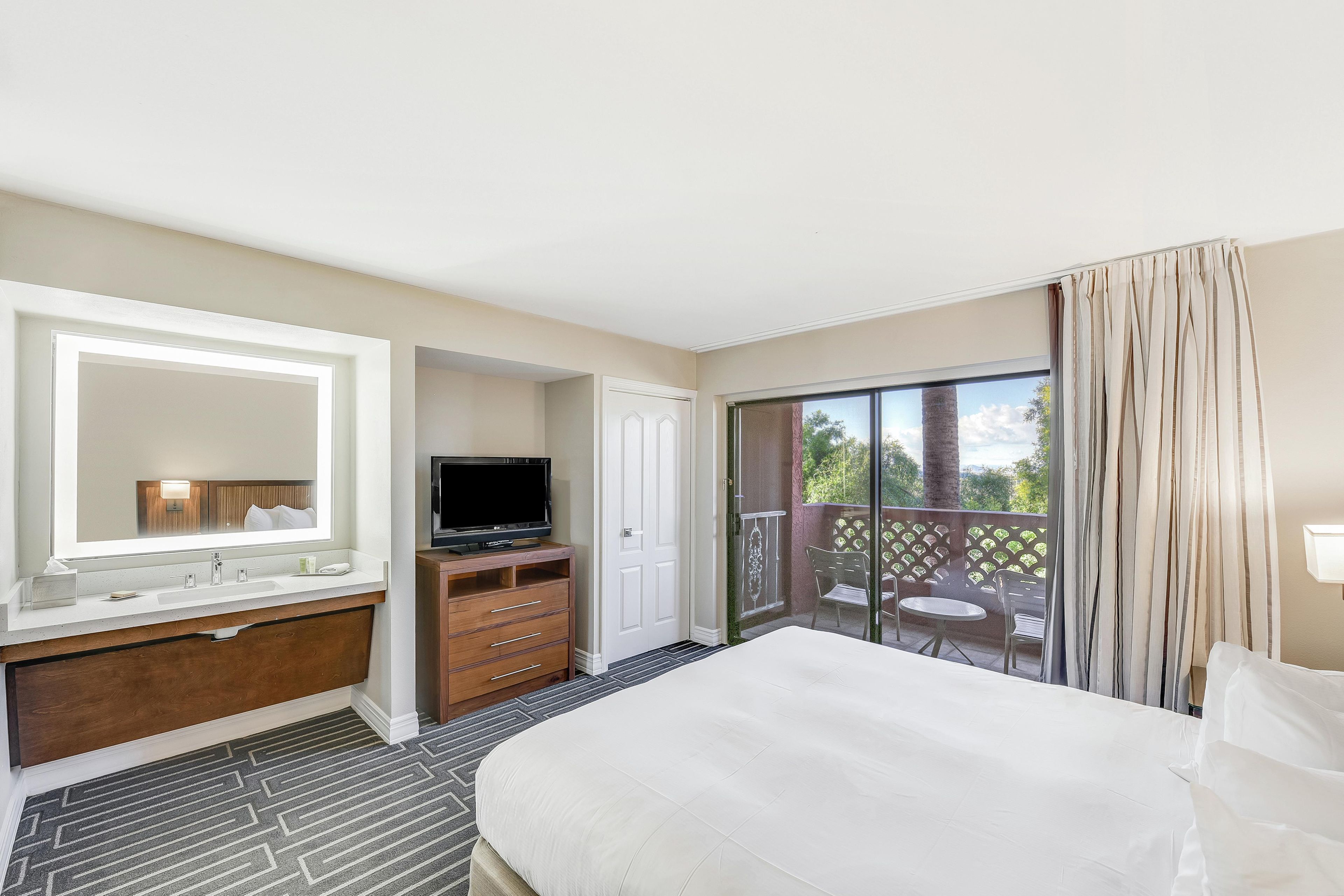 Habitación de hotel con una cama grande, baño con tocador y espejo, televisor y balcón con mesa y sillas.