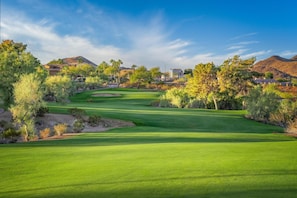 A golf course with a clear sky, surrounded by trees and distant hills.