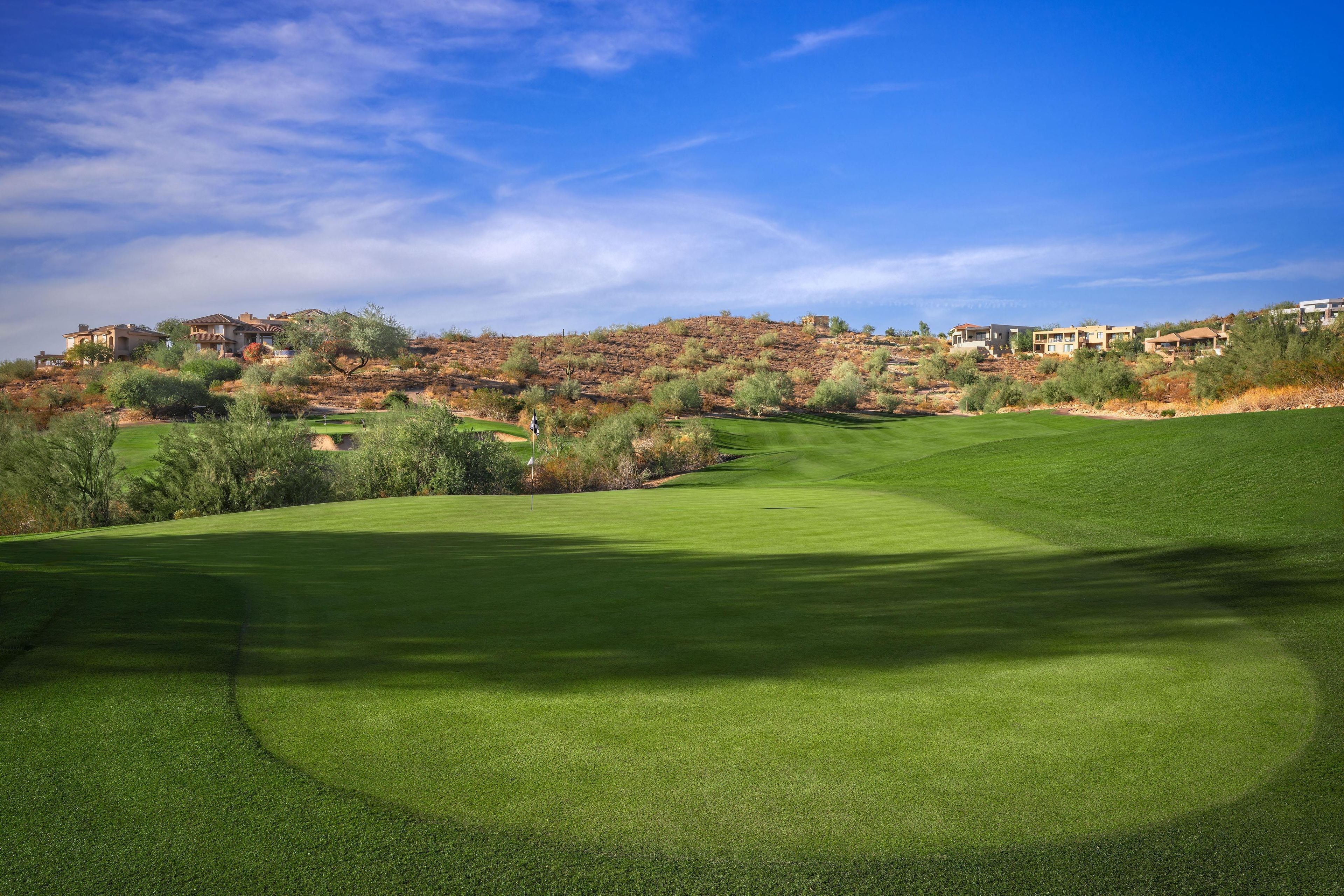 A golf course with well-maintained grass, a clear sky, and a hilly landscape with buildings in the background.