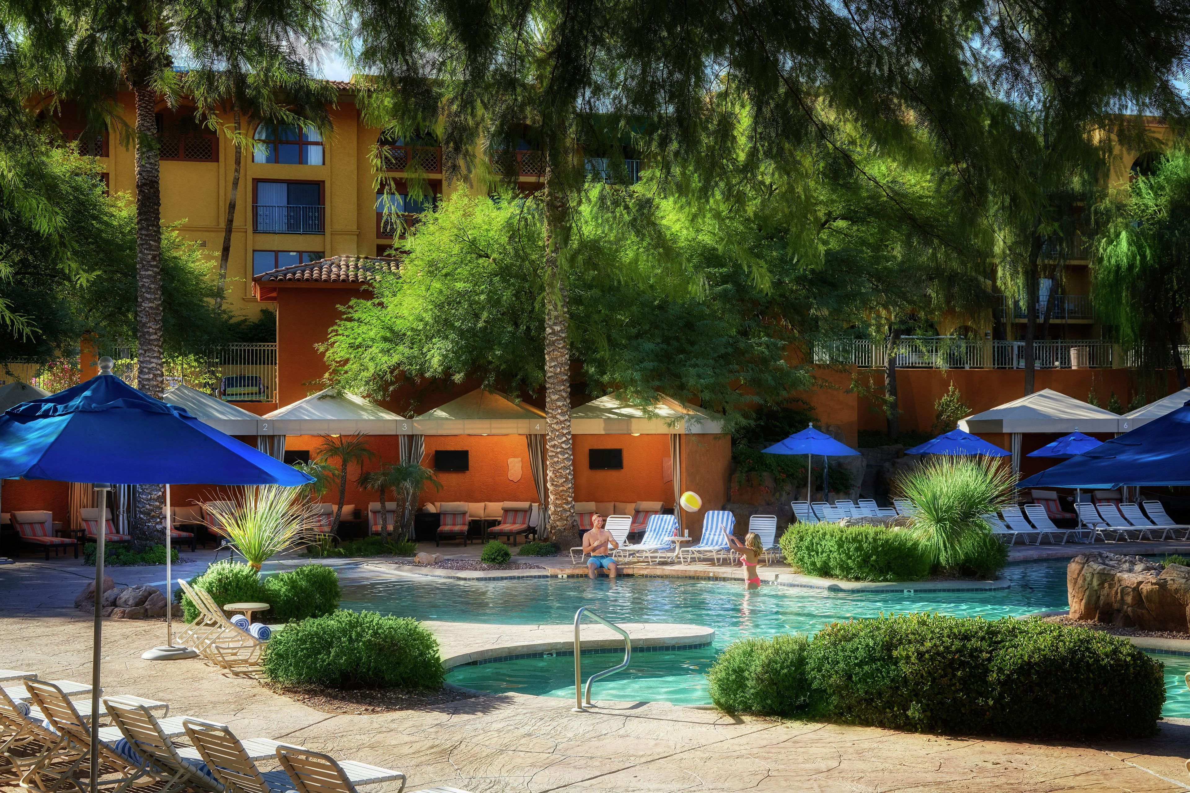 Un área de piscina con sillones de descanso y sombrillas, rodeada de vegetación y un edificio con balcones.