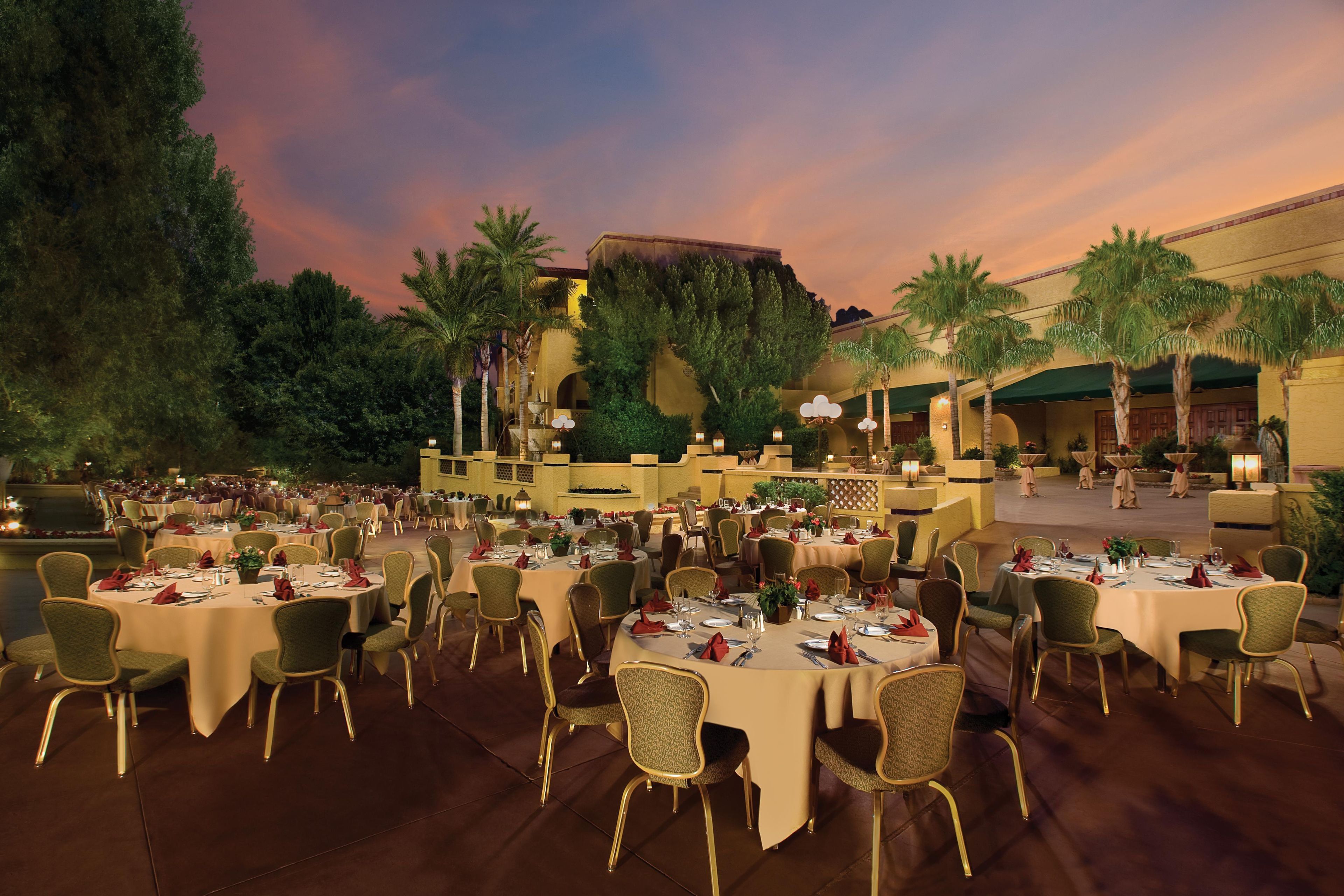 An outdoor dining area with round tables and chairs, surrounded by palm trees and a building with arches.