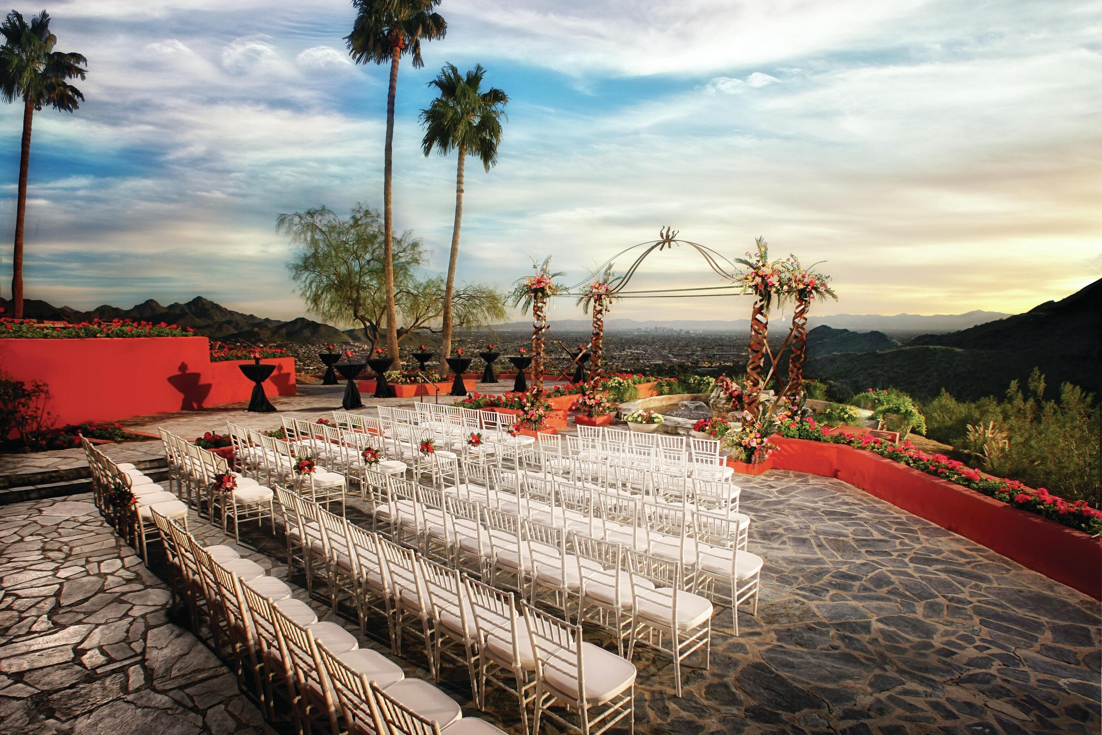 An outdoor event space with rows of white chairs and tables, palm trees, and a mountainous backdrop.