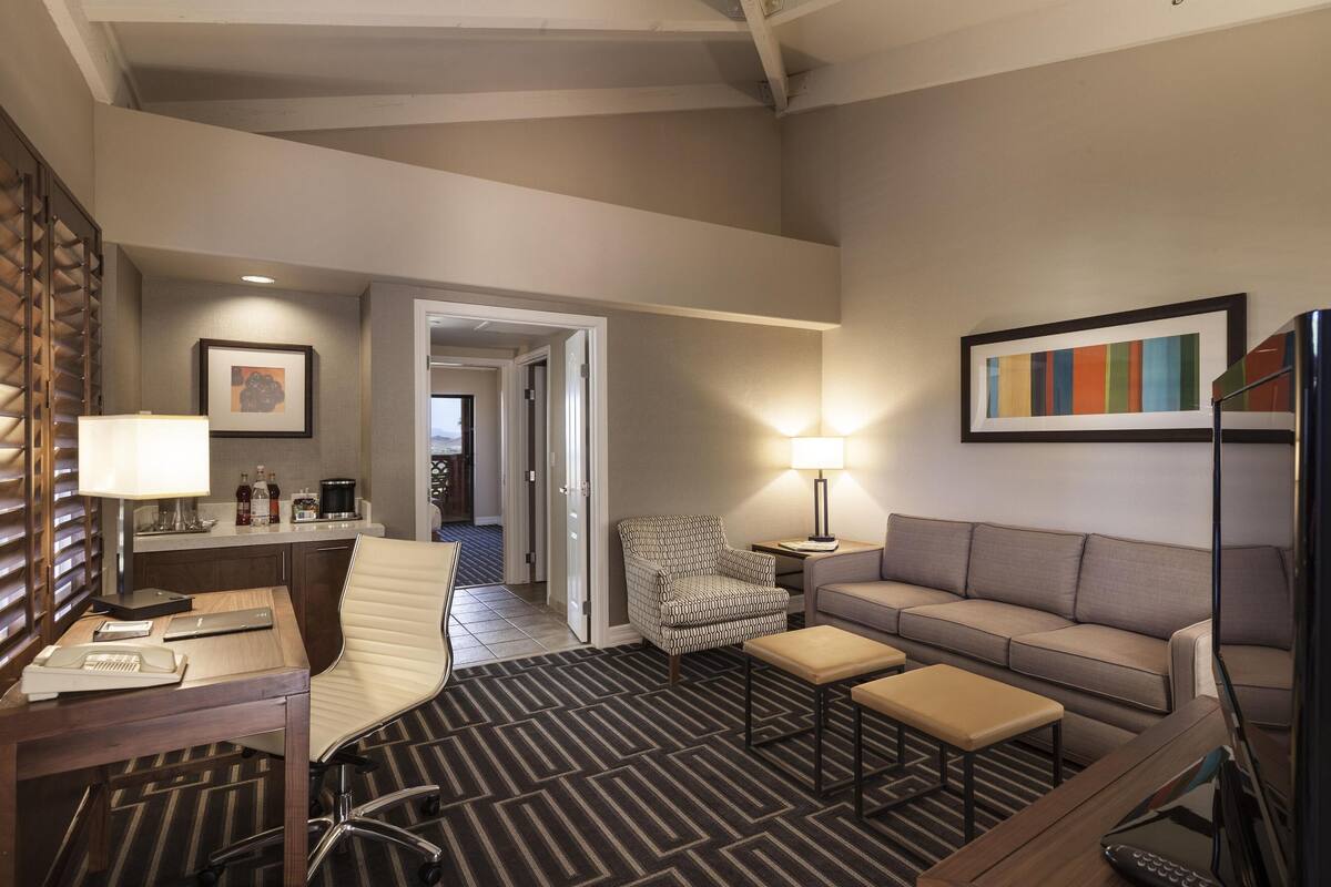 Habitación de hotel con escritorio, silla, sofá y televisor. Se observa un cuadro en la pared y una lámpara sobre el escritorio.