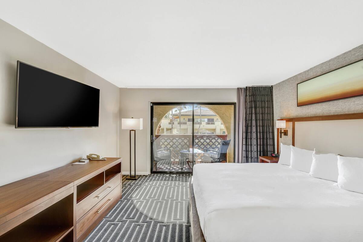 Habitación de hotel con cama, escritorio, televisión, balcón con vistas y puerta.