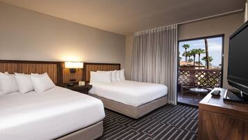 A hotel room with two beds, a TV, a desk, and a balcony with a view of palm trees.