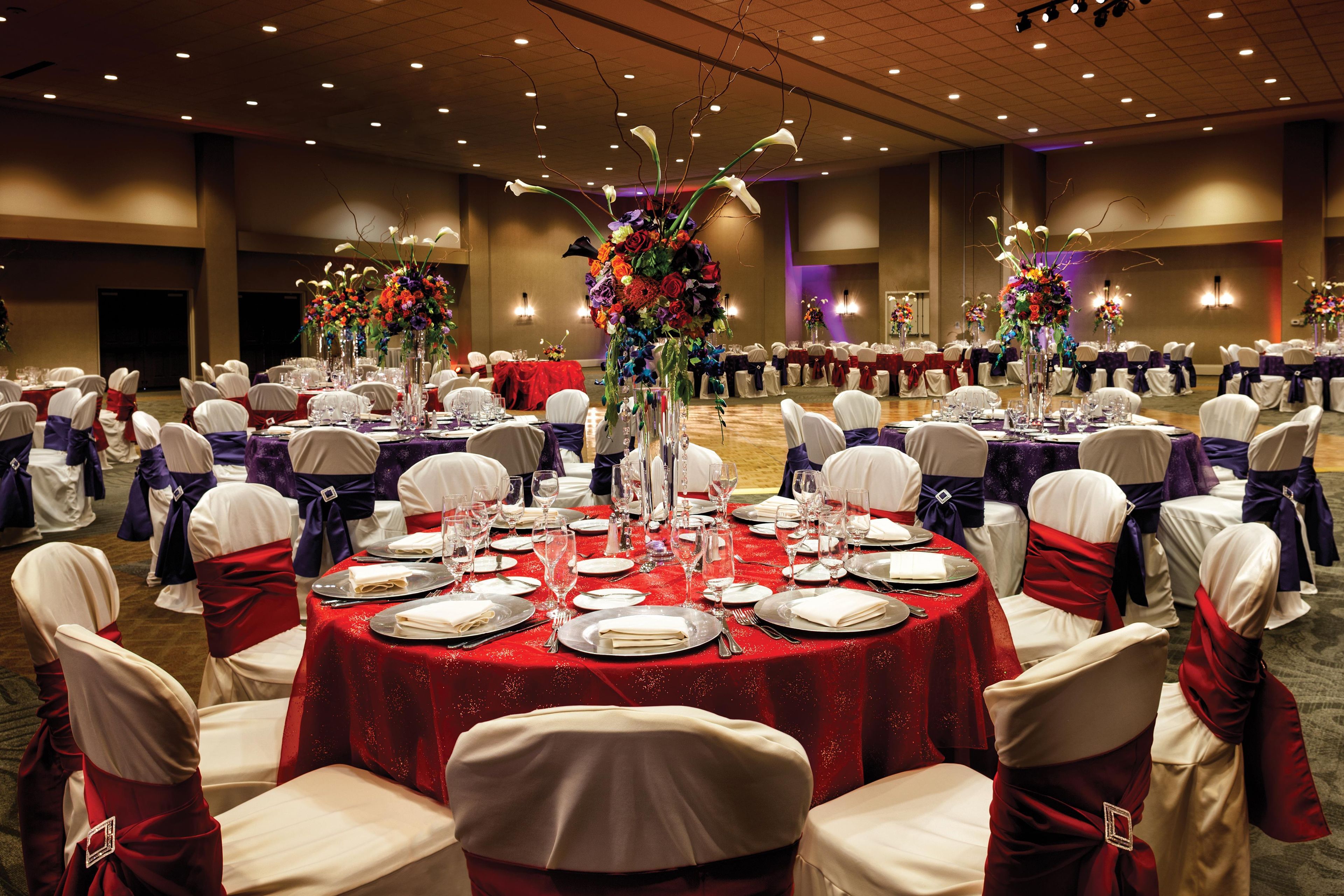 Un salón de banquetes con mesas redondas dispuestas para cenar, sillas blancas con cintas rojas y centros de mesa florales.