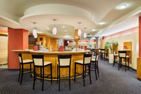 Un hall de hotel con una zona de bar central, varios conjuntos de asientos y una cocina al fondo.