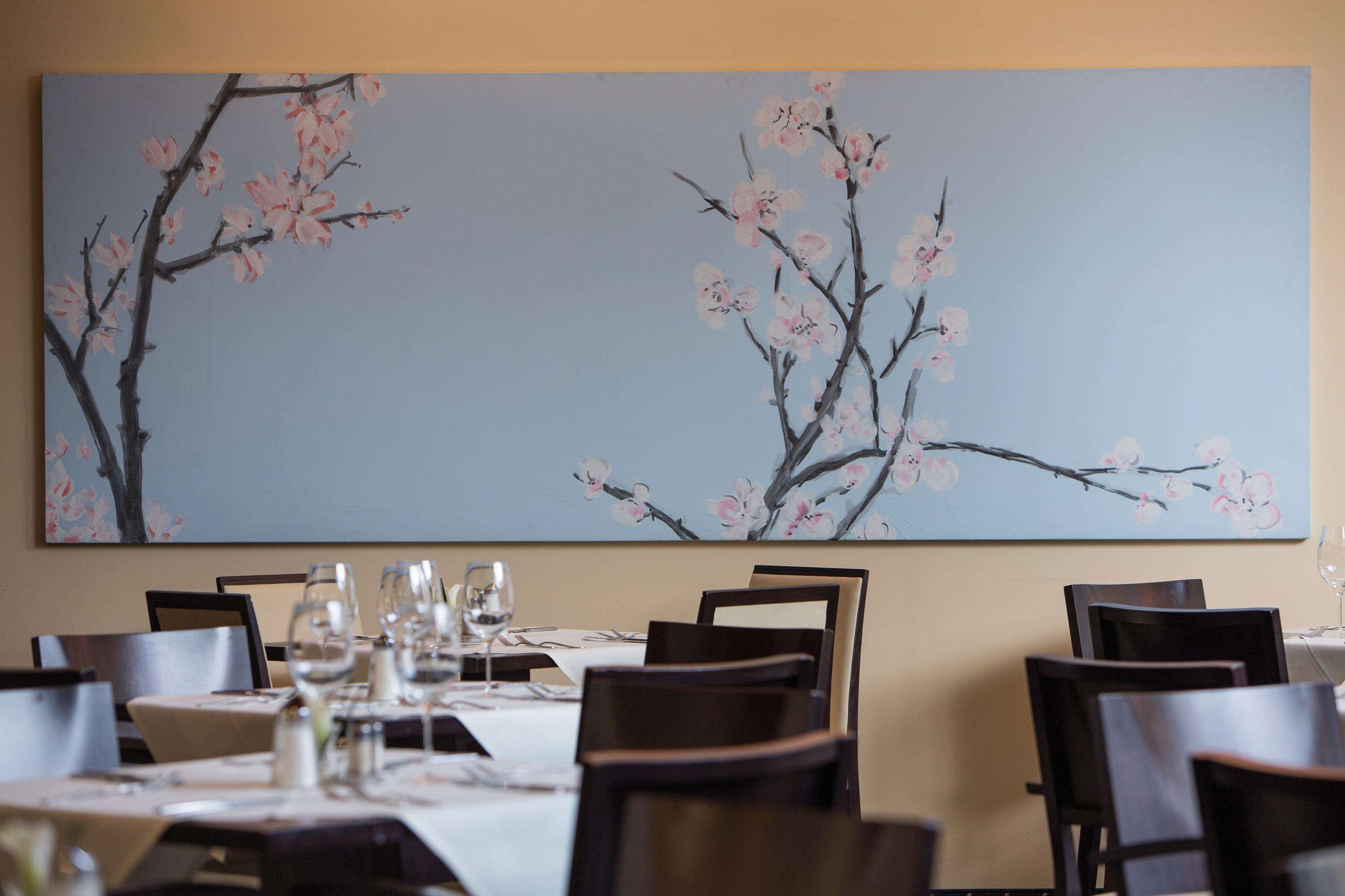 A dining area with tables set for a meal, chairs, and a large painting of cherry blossoms on the wall.