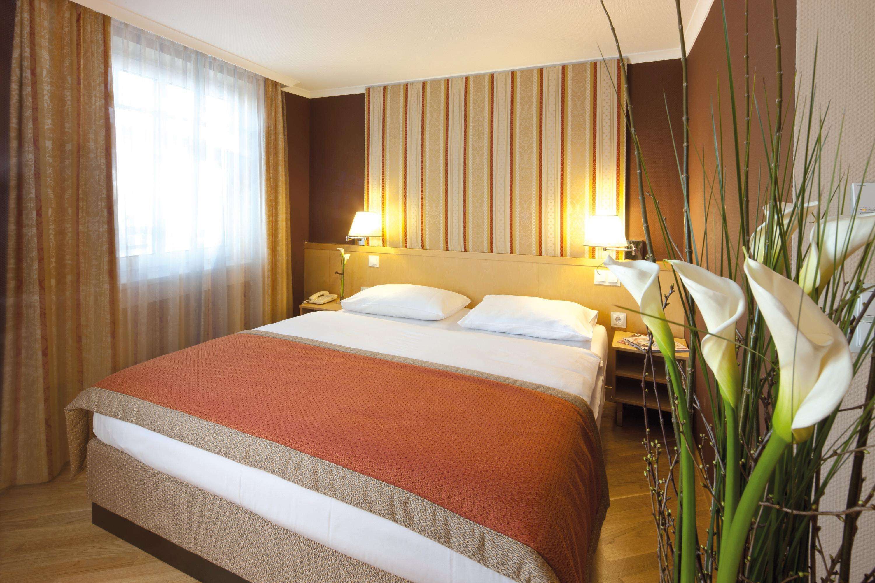 A hotel room with a large bed, two bedside tables, a nightstand, and a lamp. There is a window with curtains, a wall with vertical stripes, and a plant with white flowers.