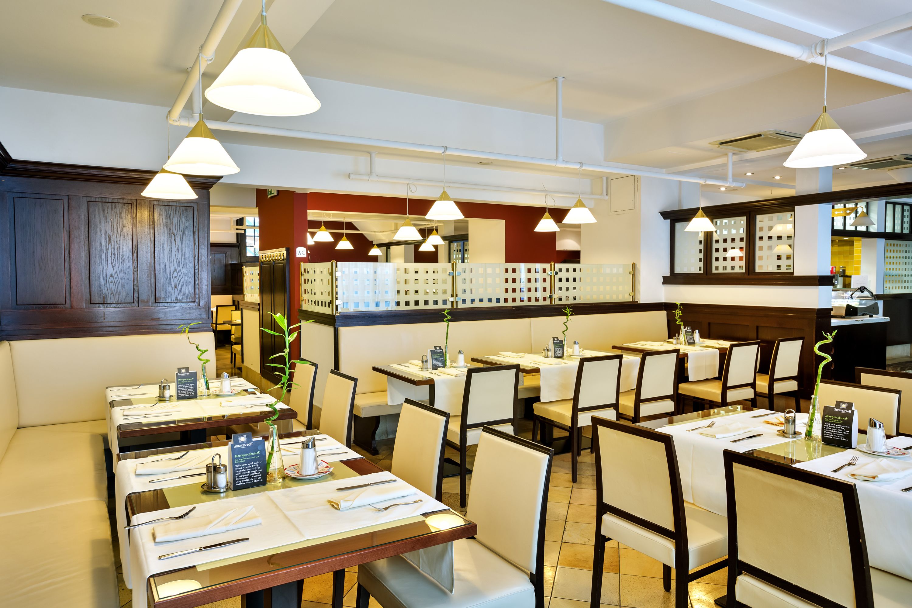 Interior de restaurante com várias mesas, cadeiras, iluminação pendente e uma área de cozinha ao fundo.