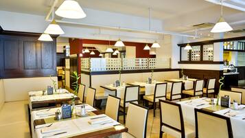 Interior de restaurante com várias mesas, cadeiras, iluminação pendente e uma área de cozinha ao fundo.