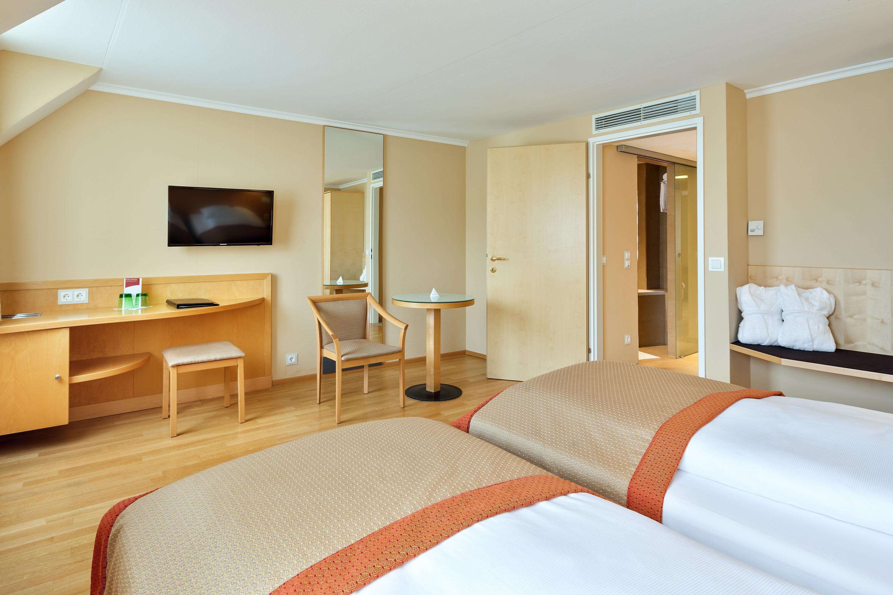 Quarto de hotel com duas camas, uma escrivaninha, uma cadeira, uma televisão e um espelho.