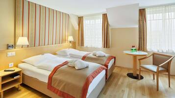 Quarto de hotel com duas camas, uma mesa pequena, uma cadeira e uma janela grande com cortinas.